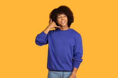 Neşeli genç bir adam canlı sarı bir arka plana karşı duruyor, sağ eliyle şakacı bir telefon konuşması yapıyor. Mavi bir kazak giyiyor ve kıvırcık saçları var.