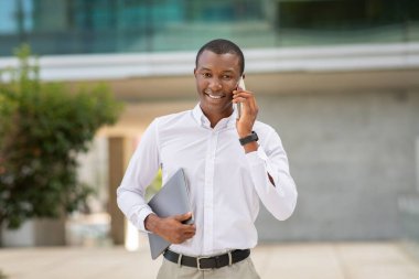Genç siyahi bir profesyonel, çağdaş bir ofis binasının önünde dururken cep telefonuyla sohbet ediyor. Bir dizüstü bilgisayar taşıyor ve kendine güveniyor.