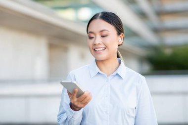 Genç bir Asyalı kadın dışarıda akıllı telefonuna bakarken gülümsüyor. Kablosuz kulaklık takıyor, bir konuşmayla meşgul olduğunu ya da müzik dinlediğini gösteriyor.