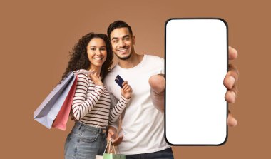 Alışveriş torbaları ve beyaz ekranlı akıllı telefon tutan mutlu bir çift. Adam telefonu sağ eliyle önünde tutarken, kadın elinde bir kredi kartı tutuyor ve gülümsüyor.