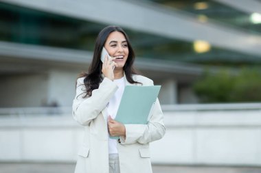 Beyaz ceketli ve beyaz gömlekli genç bir iş kadını cep telefonuyla konuşurken modern bir ofis binasının önünde duruyor.