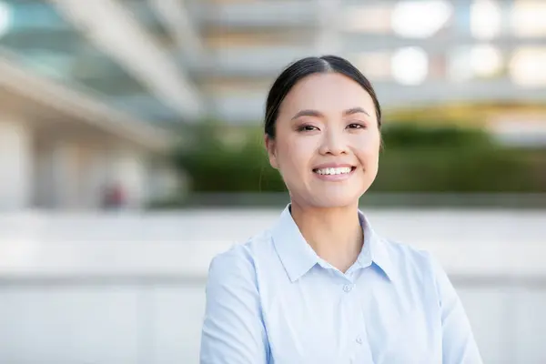 stock image A young Asian woman stands confidently outdoors, smiling warmly while wearing a light blue blouse. The setting features modern architecture and greenery in the background