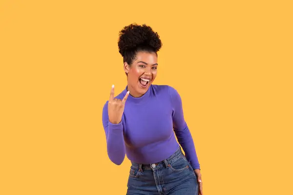 stock image A young woman stands confidently, flashing a rock hand gesture with a big smile on her face. She is wearing a fitted purple sweater paired with high-waisted jeans.