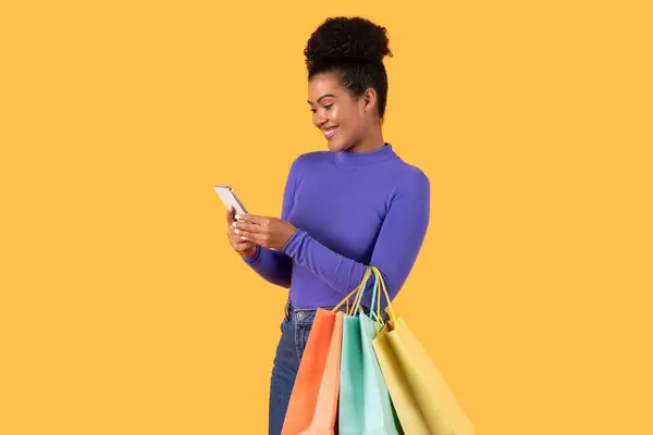 stock image A cheerful woman stands holding several vibrant shopping bags in one hand while using her smartphone with the other. She appears to be engaged with her device, showcasing a modern shopping