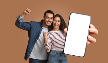 Bu görüntü, kahverengi bir arka planın önünde duran orta doğulu çifti gösteriyor. Gülümsüyorlar ve heyecanlılar, yumrukları havada. Kadın elinde boş ekranlı bir akıllı telefon tutuyor..