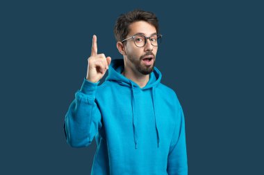 Mavi kapüşonlu genç bir adam şaşırmış ya da heyecanlı görünürken sağ işaret parmağıyla yukarı bakıyor. Gözlükleri, heyecanını yansıtan ifadesine entelektüel bir dokunuş katıyor.