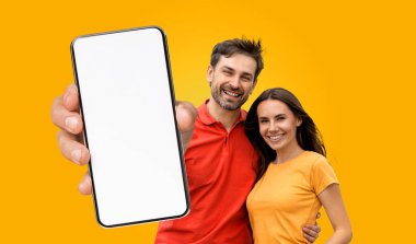 Genç bir çift kameraya gülümsüyor. Elinde parlak sarı bir arkaplan önünde boş bir ekranla akıllı bir telefon tutuyor..