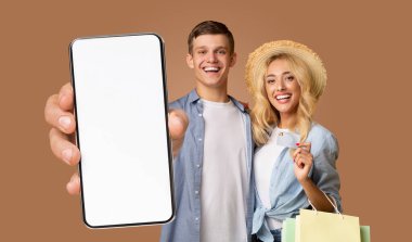 Kahverengi arka planda gülümseyen genç bir çift. Adam mavi bir ekose gömlek giyiyor ve elinde kameraya doğru beyaz bir ekran olan bir akıllı telefon tutuyor.