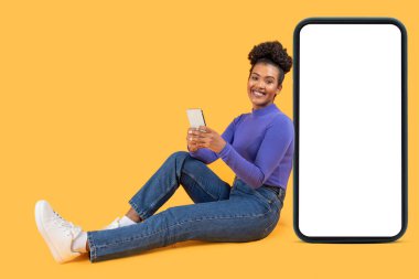 Genç bir kadın elinde akıllı bir telefonla rahatça yerde oturuyor ve mutlu bir şekilde gülümsüyor. Parlak sarı arka plan canlı bir atmosfer yaratır.