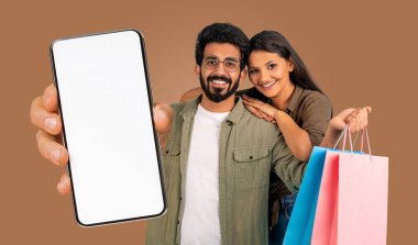 Kahverengi arka planda duran Hintli çift. Adam kameraya doğru beyaz ekranlı bir akıllı telefon tutuyor. Kadın alışveriş torbalarını tutuyor ve gülümsüyor..