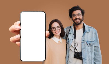 Genç, çok ırklı bir çift, kahverengi bir arkaplan önünde birlikte poz veriyor. Önlerinde boş beyaz ekranlı siyah bir akıllı telefon tutuyorlar, hem gülümsüyorlar hem de kameraya bakıyorlar.