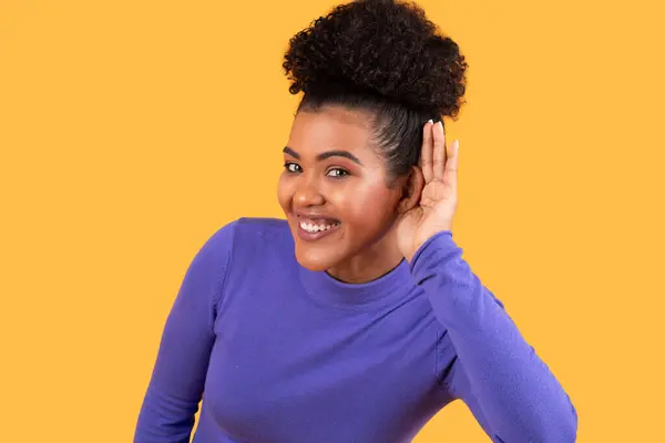 stock image A cheerful young woman raises her hand to her ear, displaying a friendly smile. She is positioned against a vibrant yellow background, creating a lively and engaging atmosphere.