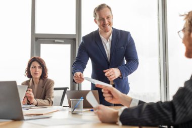 Modern bir ofiste mavi takım elbiseli bir adam masanın önünde duruyor. O, meslektaşlarına bir belge sunarken gülümser. Bir meslektaşım masada oturup adama bakıyor..