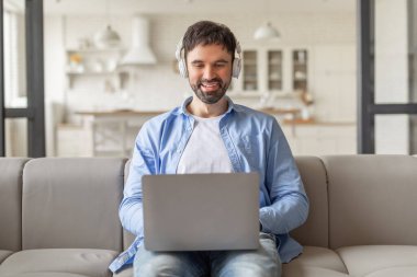 Modern bir oturma odasındaki açık gri koltukta oturan bir adam dizüstü bilgisayarı üzerinde çalışırken gülümsüyor. Kulaklık ve açık mavi düğmeli gömlek giyiyor..