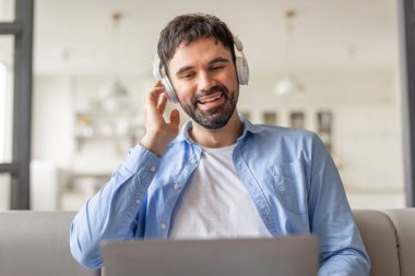Kulaklık takan bir adam evinde bir kanepede oturup müzik dinlerken gülümsüyor. Önünde açık olan bir dizüstü bilgisayarla çalışıyor..