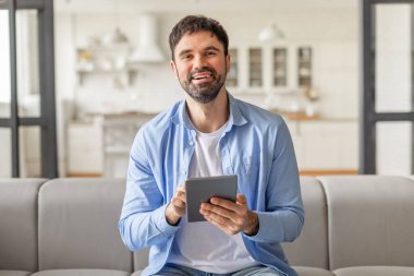 Evindeki gri bir koltukta oturan bir adam elinde tablet bilgisayar tutarken gülümsüyor, internette geziniyor ya da kayıyor.