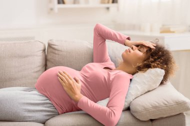 Baş ağrısı çeken genç bir kadın kanepeye uzanmış, karnına ve alnına dokunuyor.