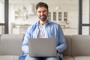 Modern bir oturma odasında açık gri bir koltukta oturan bir adam dizüstü bilgisayarıyla çalışıyor. Gülümsüyor ve işini seviyor gibi görünüyor..