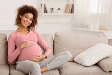 Maternity concept. Happy pregnant woman sitting on sofa and caressing her belly, empty space