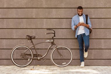 Genç siyahi adam akıllı telefon kullanıyor, bisikletinin yanında duruyor, gri şehir duvarına yaslanıyor, boş yer var.