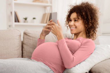 Afro-Amerikalı hamile kadın akıllı telefondan bakıyor, evdeki kanepede uzanıyor, boş yer var.