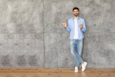 Mavi gömlekli ve kotlu bir adam gri beton bir duvara yaslanıyor, elinde bir akıllı telefon tutuyor ve sağ eliyle fotokopi alanını işaret ediyor. Gülümsüyor ve kameraya bakıyor..