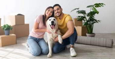 Cheery young international couple with cute golden retriever dog sitting on floor of new home on relocation day. Happy millennial husband and wife with cute pet moving to new house
