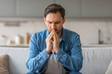 Bir adam beyaz bir kanepede oturuyor, elleri yüzünün önünde birbirine kenetlenmiş, aşağıya bakıyor, düşüncelere dalmış. Alnı çatladığı için dalgın ve muhtemelen sıkıntılı görünüyor..