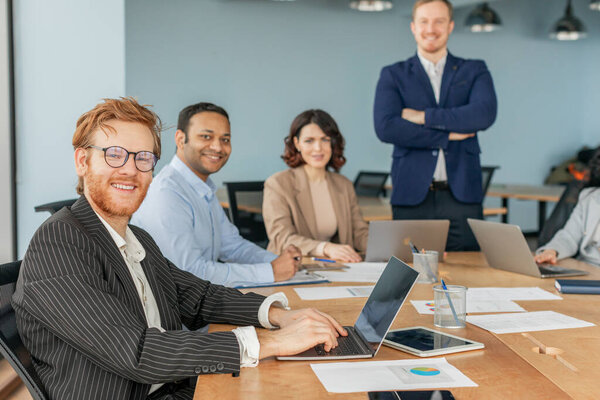 Группа из четырех профессионалов бизнеса работает вместе в современной офисной обстановке. Один сидит за столом и работает над ноутбуком, в то время как другой мужчина и женщина сидят рядом с ним