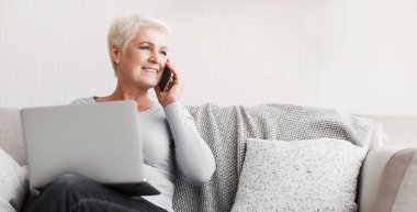 Evdeki koltukta oturan, dizüstü bilgisayarı olan ve telefonla konuşan mutlu, son sınıf öğrencisi bir kadın.