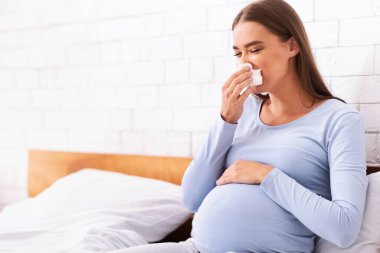 Yatak odasında yatağında oturan hasta hamile kadın hapşırıyor ve kağıt mendile burun sümkürüyor. Hamilelik ve Hastalık