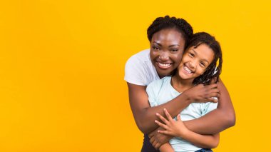 Siyah anne kızını kucaklıyor, mutlu ve çok mutlu görünüyor. Sarı stüdyo arkaplanı, kopyalama alanı
