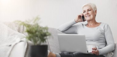 Aktif gülümseyen yaşlı kadın evdeki koltukta oturuyor, dizüstü bilgisayarını tutuyor ve telefonla konuşuyor, çay içiyor, fotokopi çekiyor.