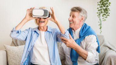 Modern teknolojiler. Olgun bir kadın, sevgi dolu kocası yakınlarda otururken VR gözlüklerini test ediyor.