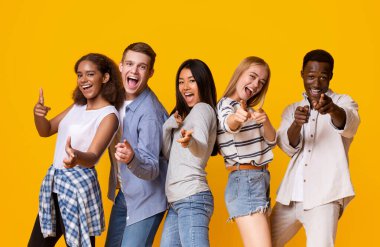 Hey there. Cheerful group of international students indicating at camera over yellow background, pointing with fingers