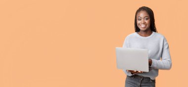 Neşeli genç siyah bir kadın canlı turuncu bir arkaplan önünde dizüstü bilgisayarını güvenle tutuyor. Pozitif ve heyecan yayıyor. Gelişigüzel bir ortamda teknolojiyle meşgul oluyor..