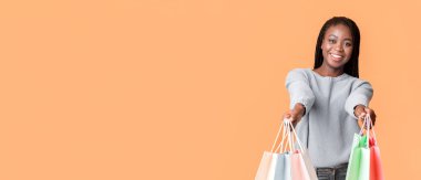 Genç bir Afrikalı Amerikalı kadın elinde birkaç renkli alışveriş çantası tutarken parlak bir şekilde gülümsüyor. Canlı turuncu bir arkaplan karşısında kendinden emin duruyor, neşe ve heyecan saçıyor, boşluğu kopyalıyor.