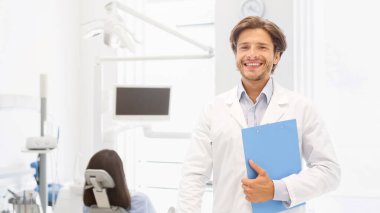 Kliniğimi ziyaret edin. Yakışıklı dişçi yüzünde gülücükler açıyor ve klasörü arka planda, boş alanda tutuyor.