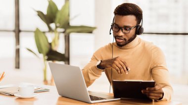 Ciddi bir Afrikalı Amerikalı destek operatörü, kulaklıklı dizüstü bilgisayara bakıyor, belge yazıyor. Boş boşluk