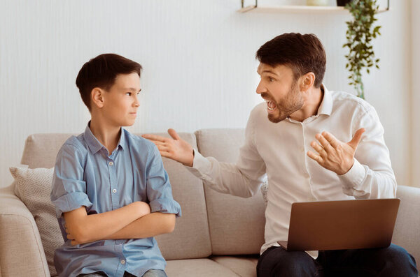 Агрессивный отец. Злой отец кричит на оскорбленного сына, работающего на ноутбуке, сидящего на диване в помещении. Селективный фокус