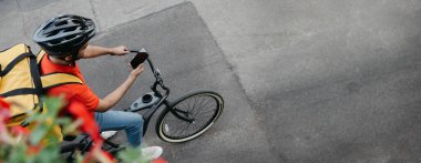 Adresi bul. Bisikletli kurye koruyucu kasklı, sırt çantalı bisikletli, bisiklete binmiş, telefona bakıyor ve caddede, üst görünümde, boş alanda online harita kullanıyor.