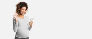 Mutlu hamile kadın cep telefonuyla video görüşmesi yapıyor ve beyaz arka planda el sallıyor.