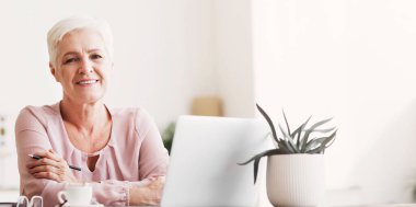 Evde çalışma masasında poz veren çekici yaşlı kadın laptopla çalışıyor, kahve içiyor, fotokopi çekiyor.