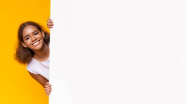 Joyful black girl looking from big white board for advertising, yellow background, panorama