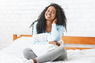 Genç Afrikalı neşeli genç bir kadının hayat tarzı portresi. Yatakta oturuyor ve gülümsüyor, yastığa sarılıyor.