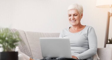 Evde çalışan gülümseyen yaşlı kadın, dizüstü bilgisayarda e-posta yazan, koltukta oturan, fotokopi çeken.