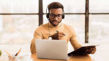 Konferans Görüşmesi. Kulaklık takmış siyahi bir adam görüntülü konuşmada notlar alıyor, müşteri danışmanlığı yapıyor.