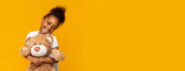 Neşeli genç siyah bir kız büyük bir oyuncak ayıya sarılırken geniş bir gülümseme sergiliyor. Canlı sarı bir arkaplan karşısında duruyor. Sevdiği oyuncağa olan ilgisini ve heyecanını gösteriyor. Kopya alanı.