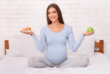 Elma tutan hamile kadın ve kruvasan evde yatakta oturarak sağlıklı bir seçim yapıyor. Hamilelikte Beslenme