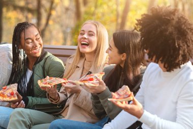 Neşeli çokkültürlü gençler şaka yapıyor ve gülüyorlar pizza yerken, parkta bankta otururken